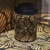 A black metal lantern with a leaf pattern is placed on a textured surface, casting intricate shadows. Produktbild-Vorschau
