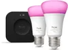A black Hue bridge and two white Hue smart bulbs with pink light are displayed against a white background. Produktbild-Vorschau