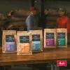 Several bags of coffee beans are displayed on a wooden table in front of two people and sacks of coffee beans. Produktbild-Vorschau