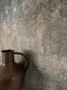 A close-up shot of a vintage-style bronze jug against a textured, weathered wall. Produktbild-Vorschau
