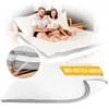 A couple is sitting on a mattress topper on a bed, reading a book. A close-up inset shows the corner of the topper being attached to a mattress. Another inset shows the underside of the topper with text. Produktbild-Vorschau