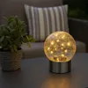 A decorative solar-powered lamp with fairy lights sits on an outdoor table next to a potted plant, with a cushion on a chair in the background. Produktbild-Vorschau