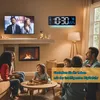 A family is gathered in a living room, watching TV and interacting with each other. A digital clock is mounted on the wall above a fireplace. Produktbild-Vorschau