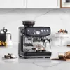 A black espresso machine with a grinder on top is shown on a kitchen counter with a cake and brownies in the background. Produktbild-Vorschau
