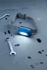 A blue Philips headlamp with its light on, placed on a gray surface with oil stains and two wrenches. Náhled obrázku produktu
