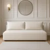 A cream-colored sofa with two large throw pillows is shown against a paneled white wall, with a round gold-framed mirror and a modern wall sconce above it. A neutral-colored rug is visible in the foreground. Produktbild-Vorschau