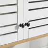 A close-up view of two black cabinet knobs on a white cabinet door with slatted panels. Náhled obrázku produktu
