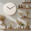 A minimalist wall clock is displayed on a light-colored wall, with several shelves filled with decorative vases and dried flowers arranged around it. Voir les images du produit