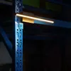 A close-up of a gold LED light bar attached to a blue metal shelf support in a warehouse or industrial setting. Produktbild-Vorschau