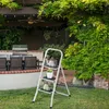 A white step stool with decorative items on it is placed on a grassy lawn in a backyard setting, with a patio, grill, and lush greenery in the background. Produktbild-Vorschau
