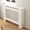 A white radiator cover with vertical slats is shown in a room with a window and wooden floor. Náhled obrázku produktu