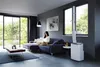 A woman is sitting on a sofa in a modern living room, using a laptop. A portable air conditioner is visible next to a window. Produktbild-Vorschau