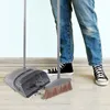 A person wearing blue jeans and black sneakers is standing on a wooden floor next to a gray broom and dustpan set. The dustpan contains some dirt. Anteprima dell’immagine prodotto
