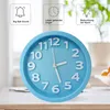 A close-up of a light blue analog alarm clock with white numbers and hands, set on a wooden surface. The background is blurred, showing hints of greenery and a white bowl. Produktbild-Vorschau
