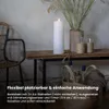 A white LED candle is placed on a wooden table next to a vase with dried grass and two glasses. Produktbild-Vorschau