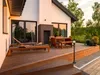 A modern house with a wooden deck and outdoor furniture, illuminated by the warm glow of sunset. Produktbild-Vorschau
