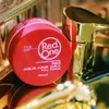 A red hair wax product is placed next to a golden faucet. Voir les images du produit