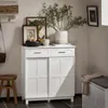 A white cabinet with two drawers and two doors, decorated with framed pictures and a vase with greenery on top, is shown in a room setting. Produktbild-Vorschau