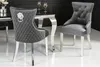 Two gray velvet dining chairs with silver legs and decorative studs are shown in a dining room setting. Produktbild-Vorschau