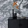 A black cabinet with fluted doors is shown in front of a black and white forest wallpaper. Two black vases and a small bowl are on top of the cabinet. Produktbild-Vorschau