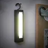 A black LED work light hangs from a hook on a textured white wall, with a wooden panel and a wheel visible on the right. Produktbild-Vorschau