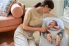 A mother is interacting with her baby in a bouncer next to a couch. Produktbild-Vorschau