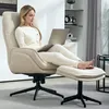 A woman is sitting in a beige armchair with a laptop on her lap, with her feet resting on a matching ottoman. The scene is set in a modern living room with shelves in the background. Voir les images du produit