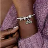 A close-up shot of a person's wrist wearing a Pandora bracelet with charms, including a bicycle charm, a horse charm, and letter charms. The person is wearing a purple knitted sweater. Anteprima dell’immagine prodotto