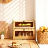 A kitchen scene with a wooden bread box, fruit, and bread on a table. Produktbild-Vorschau