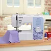 A Brother XM2701S sewing machine is shown on a wooden table with fabric and spools of thread. Voir les images du produit