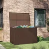A brown poly rattan garden storage box with its lid open, filled with garden tools, placed on a green lawn next to a brick wall with a window. Voir les images du produit