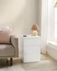 A white bedside table with three drawers is placed next to a sofa and a window. A lamp and a phone are on top of the table. Náhled obrázku produktu