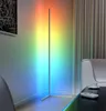 A modern living room corner with a tall, slim LED floor lamp casting a gradient of rainbow colors on the wall. A white sofa and a dark coffee table with a remote control and a cup are visible. Náhľad obrázku produktu
