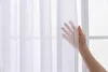 A hand is shown gently touching a white sheer curtain in front of a window. Produktbild-Vorschau