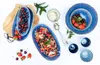 A top-down view of a fruit salad and berries served in blue stoneware dishes, with cups and spoons. Produktbild-Vorschau