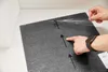 Hands are shown peeling protective film off a dark grey carpet tile, with arrows indicating a direction of application. Produktbild-Vorschau