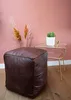 A dark brown leather pouf sits on a wooden floor next to a small glass and gold side table with a potted cactus and a small gold object. Náhled obrázku produktu