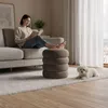 A woman is reading a book while sitting on a sofa, with her feet resting on a pouf. A small white dog is lying on the rug next to the pouf. Produktbild-Vorschau