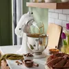 A white KitchenAid stand mixer is shown on a marble countertop, with ingredients and a bundt cake nearby, suggesting baking. Produktbild-Vorschau