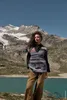 A woman stands in front of a mountain lake and snow-capped mountains, wearing a colorful knitted vest over a black shirt and brown pants. Produktbild-Vorschau