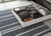 A bed with its storage compartment open, revealing pillows and boxes, placed on a rug. Produktbild-Vorschau
