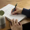 A person's hands are writing on a notebook with a blue pen. Voir les images du produit