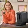 A woman is holding a cup of coffee next to a coffee machine. Produktbild-Vorschau