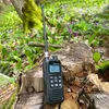 A black CB radio sits on a tree stump in a forest setting with green foliage and purple flowers. Voir les images du produit