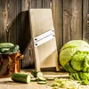 A wooden mandoline slicer stands upright on a wooden table, with various vegetables like cabbage, zucchini, and cucumbers arranged around it. Náhled obrázku produktu