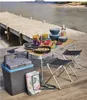A picnic setup on a wooden dock by the water, featuring a folding table laden with food and dishes, cooler, and chairs. Produktbild-Vorschau