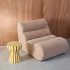 A fluffy beige armchair and a striped side table are placed in a room with wood-paneled walls. Produktbild-Vorschau
