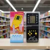 Two electronic brick game consoles are displayed on a wooden surface in front of shelves stocked with various products. Náhled obrázku produktu
