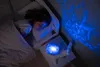 A young girl sleeps in a bed next to a whale-shaped night light that projects a blue ocean wave pattern onto the wall. Anteprima dell’immagine prodotto