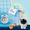 A whimsical desk setup with an astronaut pencil sharpener, a retro alarm clock, and decorative planets against a blue tiled background with a neon cloud outline. Voir les images du produit